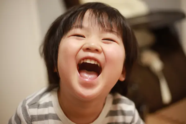 Young child with dark hair laughing with mouth wide open indoors, wearing a striped shirt.