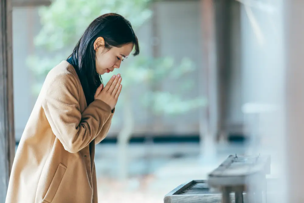 祝您旅途平安 - JAPANESE TRAVEL INSURANCE Woman in a beige coat bows with hands pressed together in prayer beside a piano-like keyboard. - JAPANESE TRAVEL INSURANCE